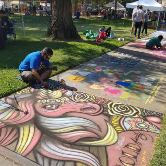 a man in a blue shirt and shorts sitting with legs crossed on the ground while working on a large colorful chalk mural on the sidewalk